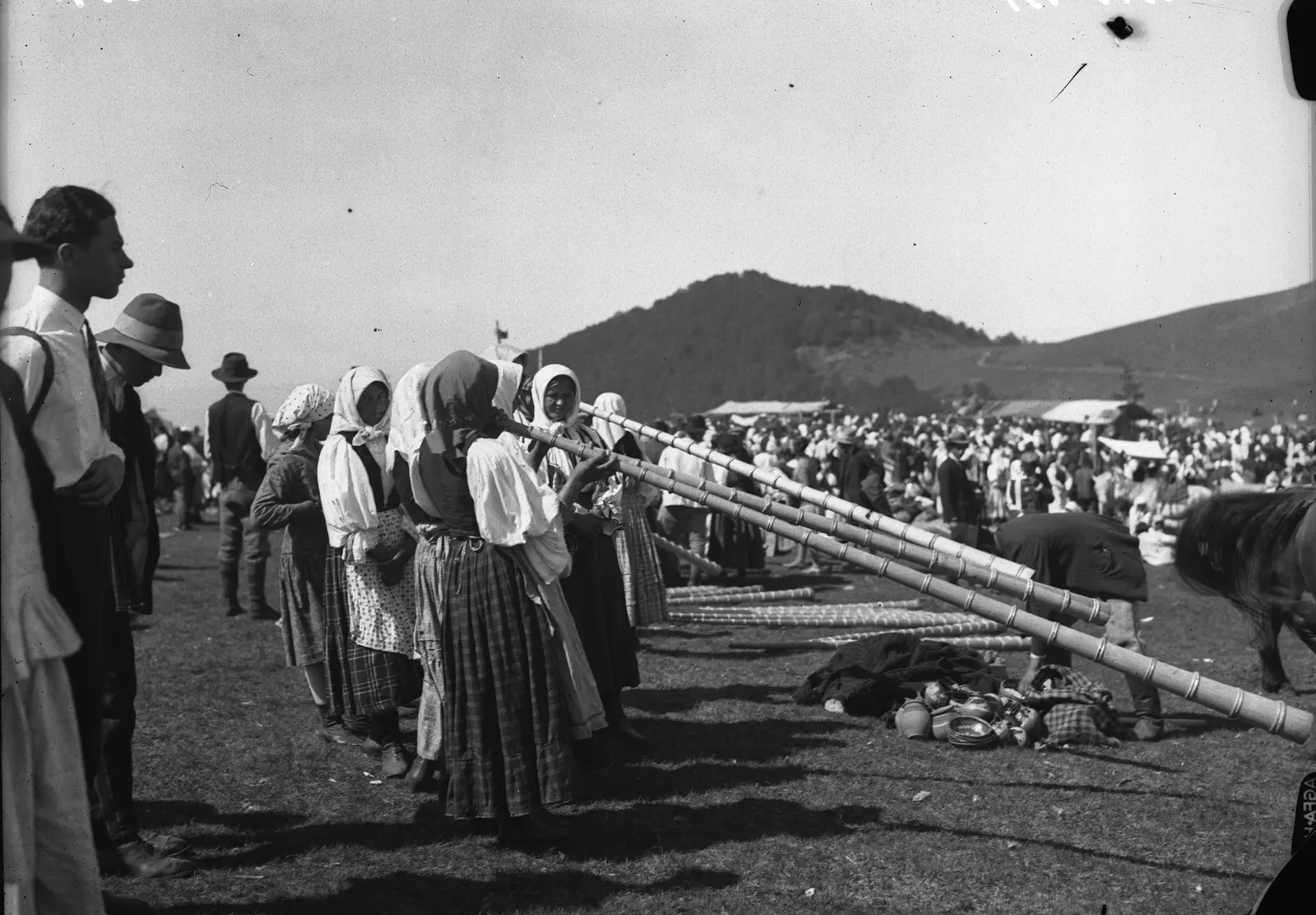 Women in headscarves play long tulnics resting on the grass, in the crowd of a mountain fair.