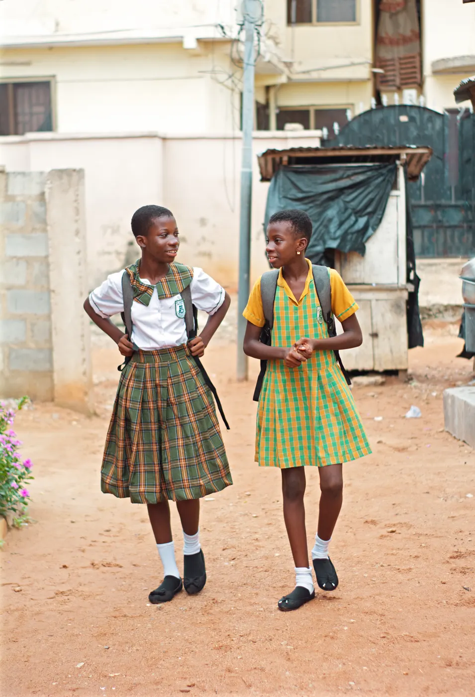 Două eleve din Accra, Ghana, 2024, în uniforme în carouri de culori diferite, cu ghiozdane în spate.