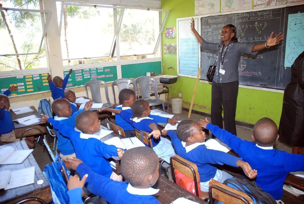 O clasă de școală primară din Kenya: elevi în pulovere albastre ridică mâna, profesorul arată spre tablă.