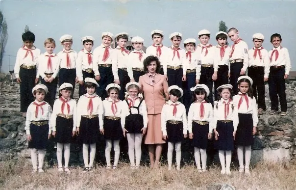 Grup de pionieri români în 1986, în cămăși albe cu cravate roșii și șepci albe, alături de o profesoară.
