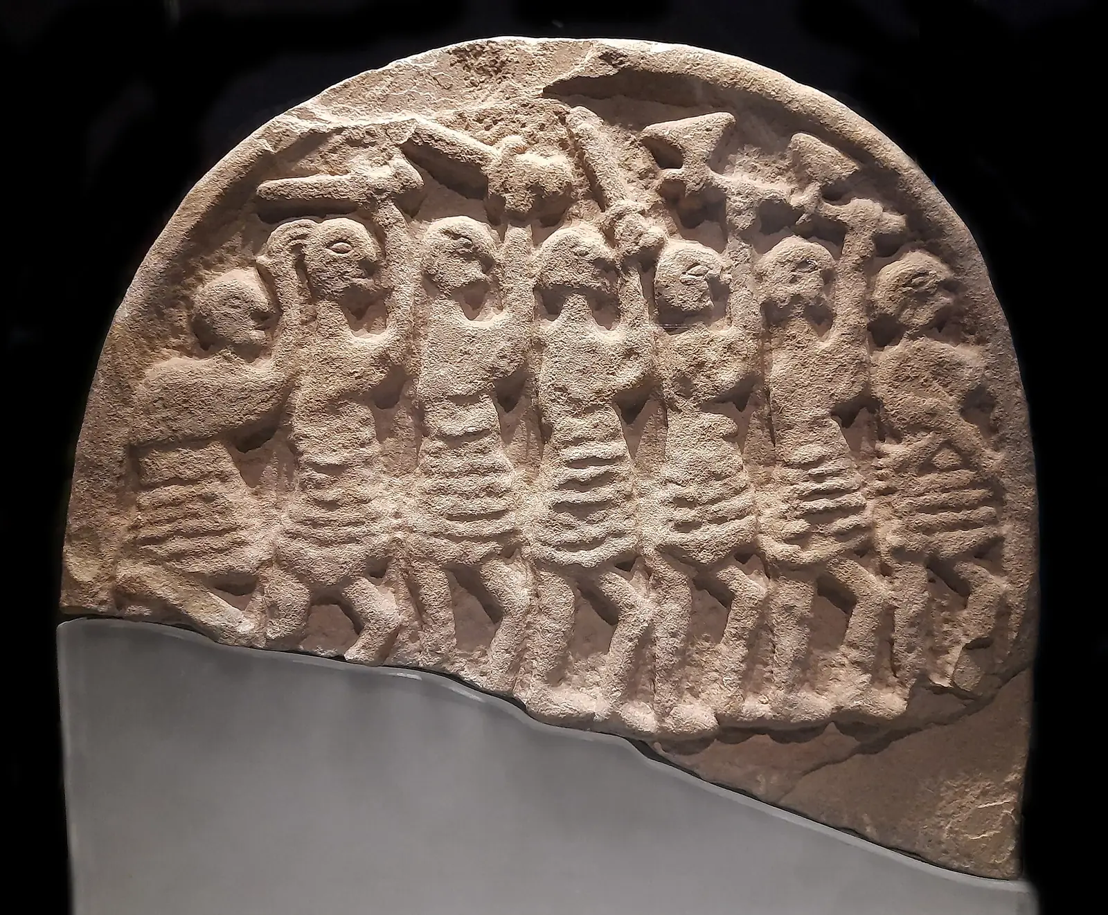 Lespede rotunjită de gresie cu șapte figuri înarmate în basorelief, cu topoare și săbii, pe fond negru.