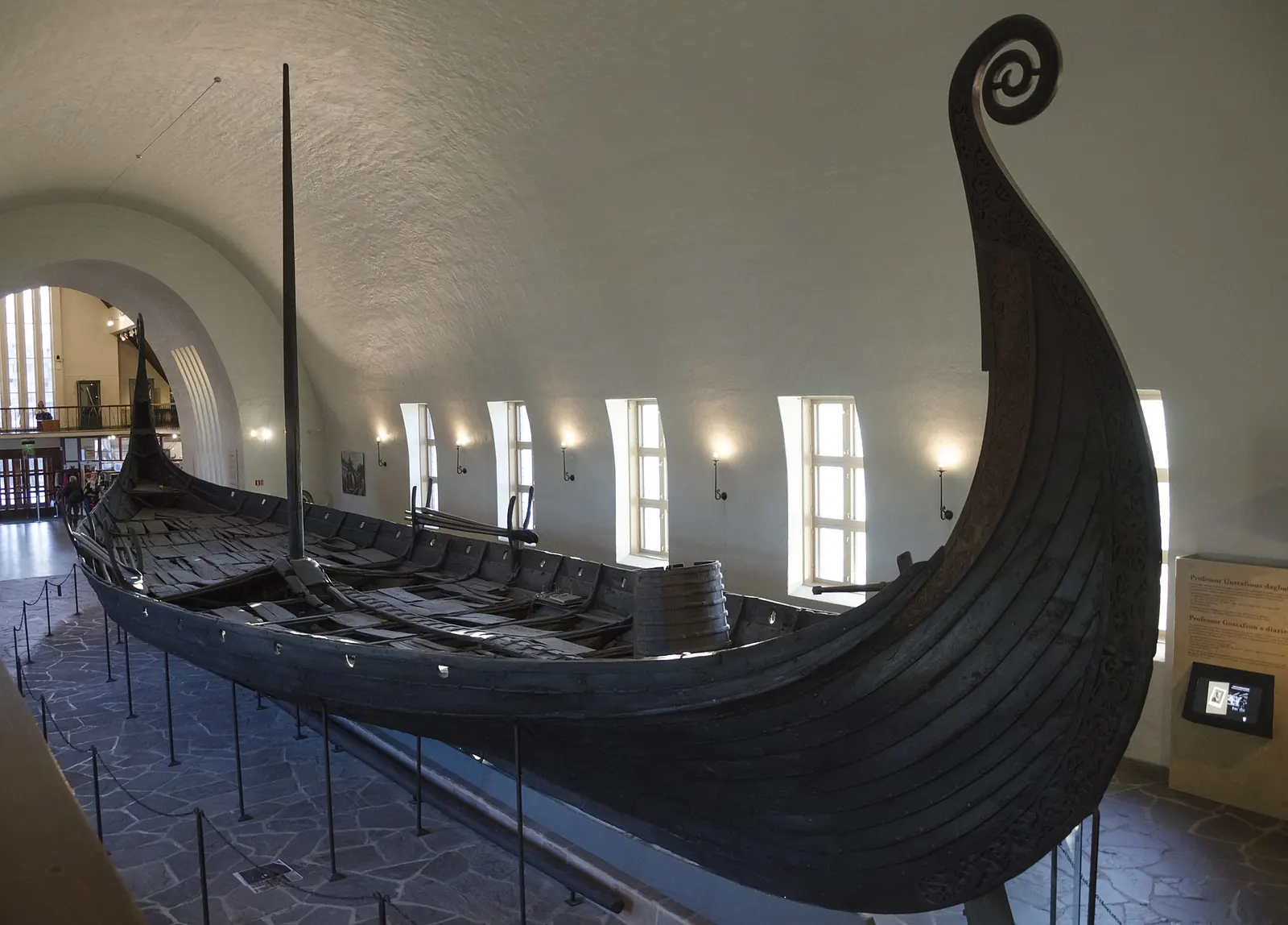 Navă vikingă întunecată, din lemn, cu prora curbată, într-o sală de muzeu albă și boltită.