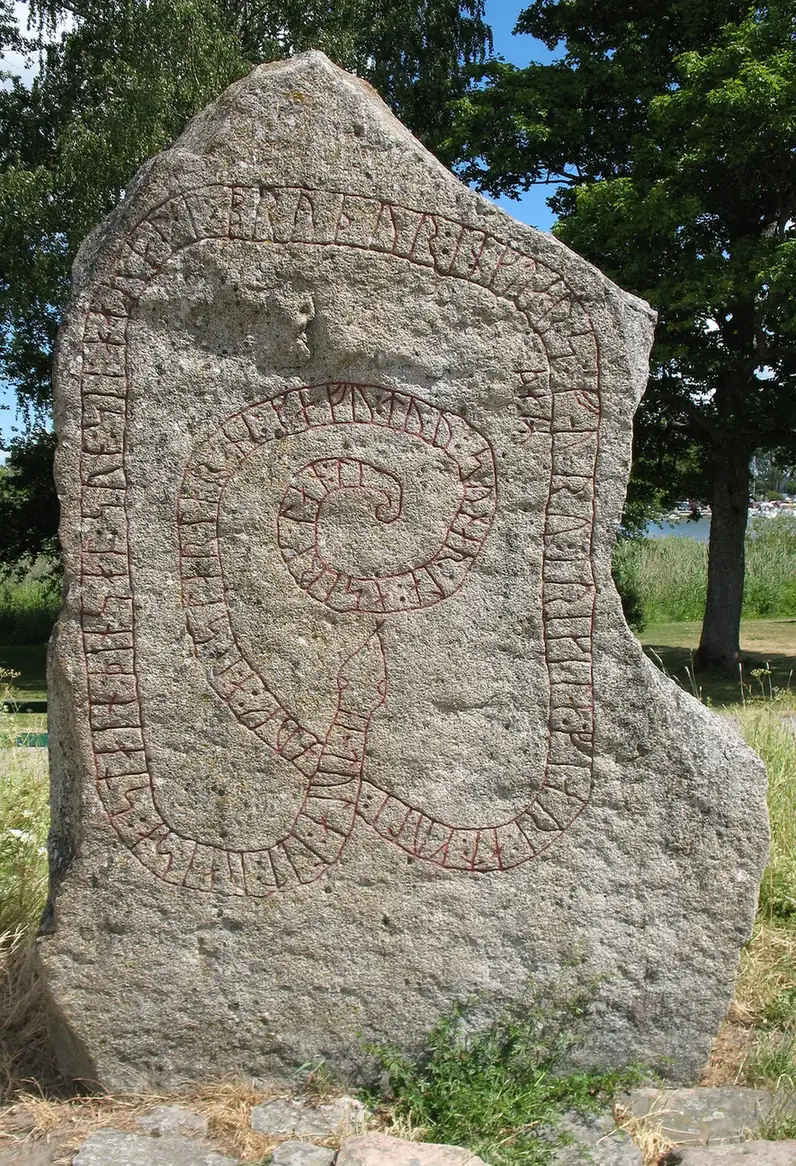 Piatră runică de granit gri, cu o bandă de rune în formă de șarpe, în iarbă, sub copaci.