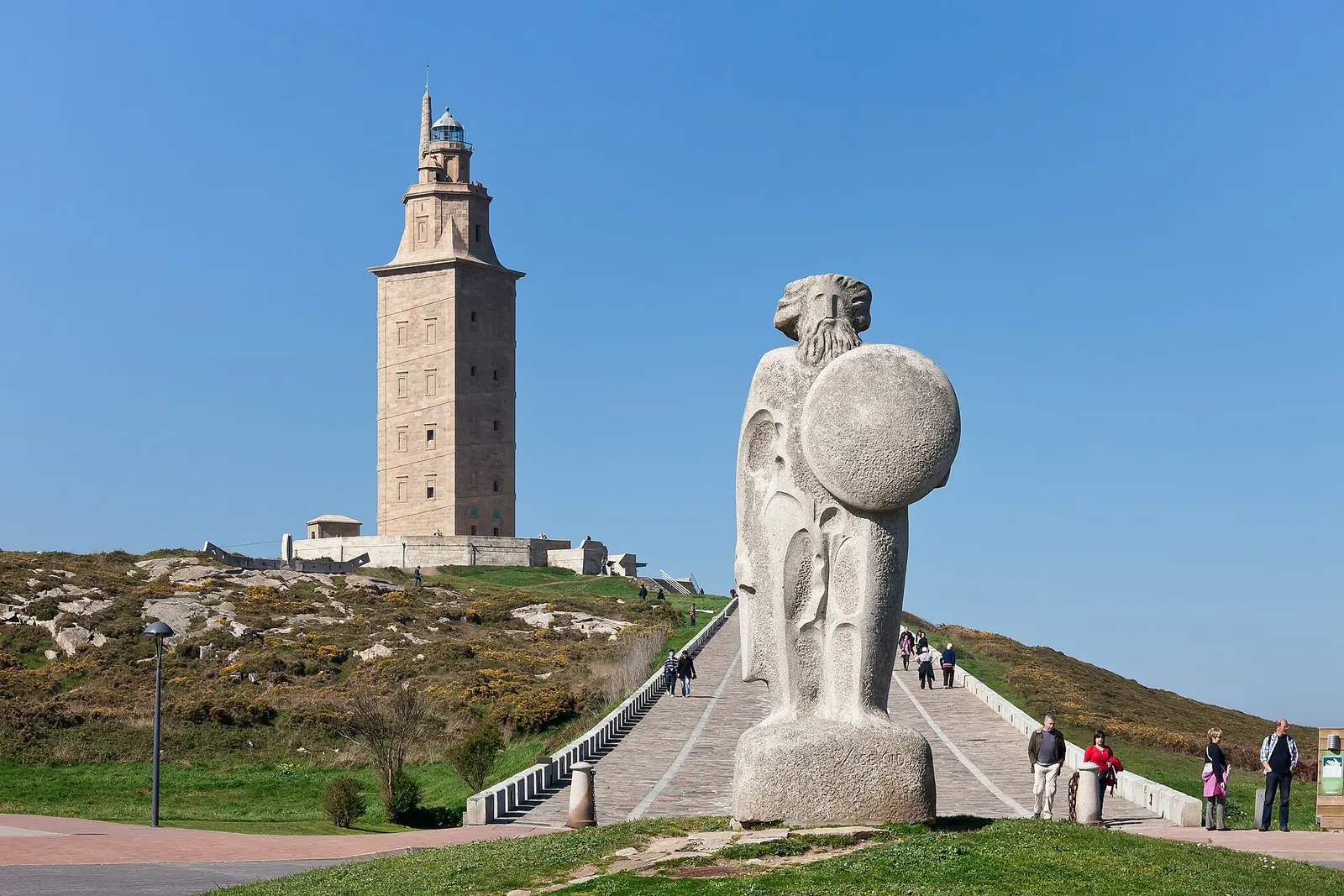 Un far înalt de piatră pe un deal, cu o statuie mare de granit a unui bărbat cu barbă și scut în prim-plan.