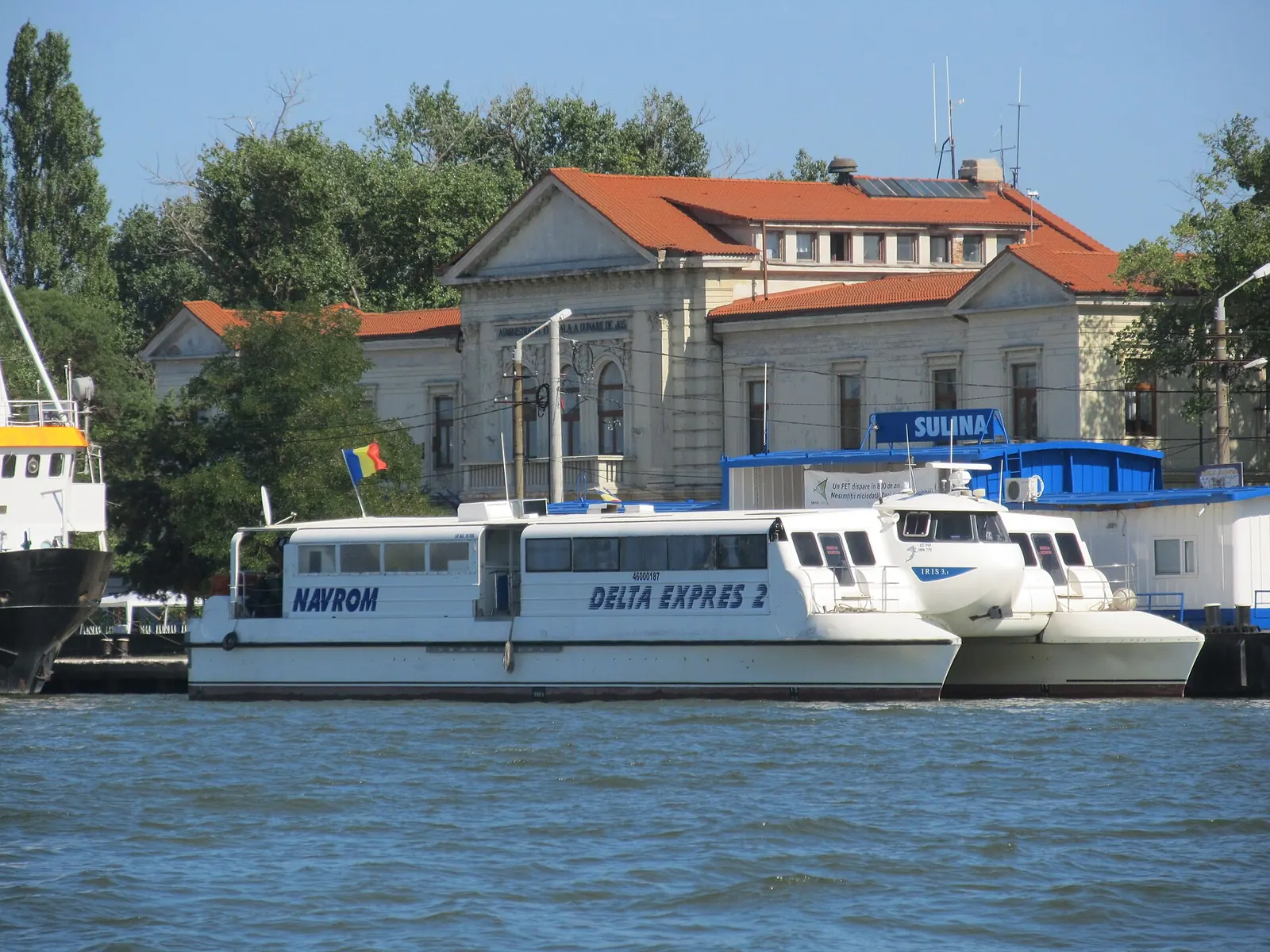 Un catamaran de pasageri acostat pe râu, în fața unei clădiri terminal cu acoperiș de țiglă roșie.