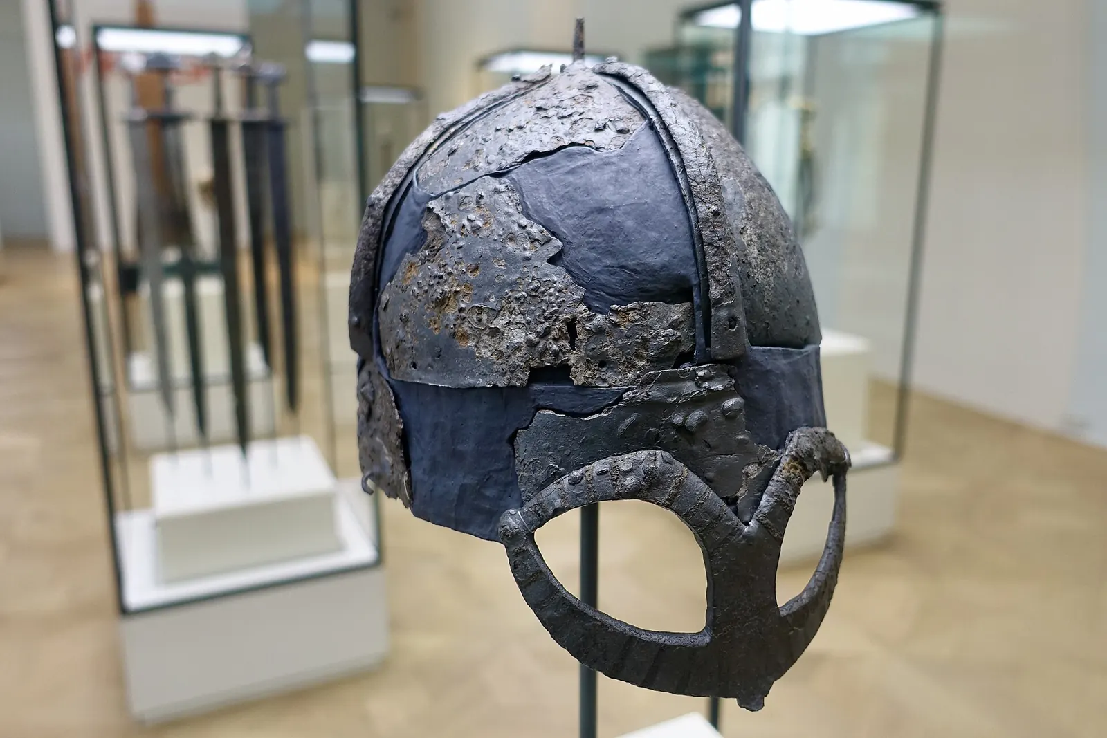 Coif de fier corodat, cu calotă rotunjită și apărătoare de ochi în formă de ochelari, într-o vitrină de muzeu.